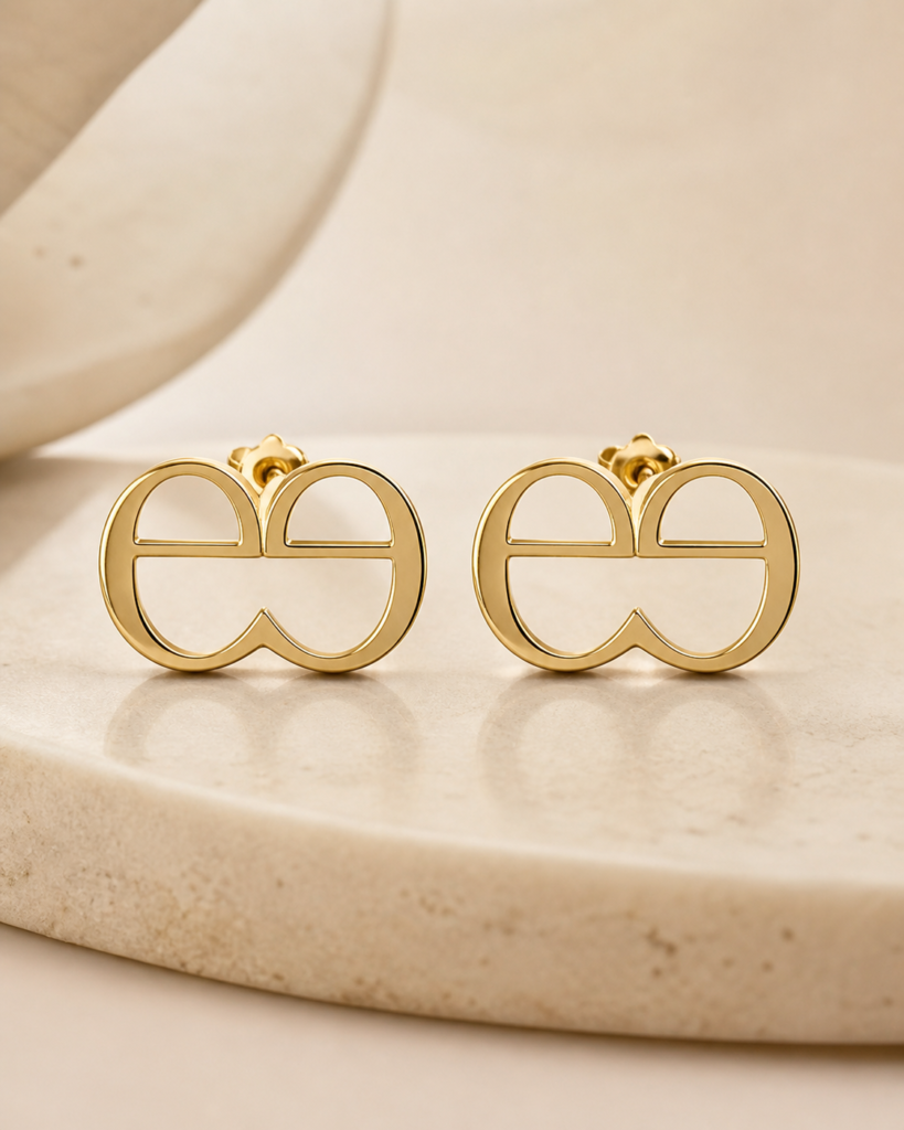 Gold deesign double-e monogram stud earrings displayed on a warm beige stone surface with soft luxury lighting.